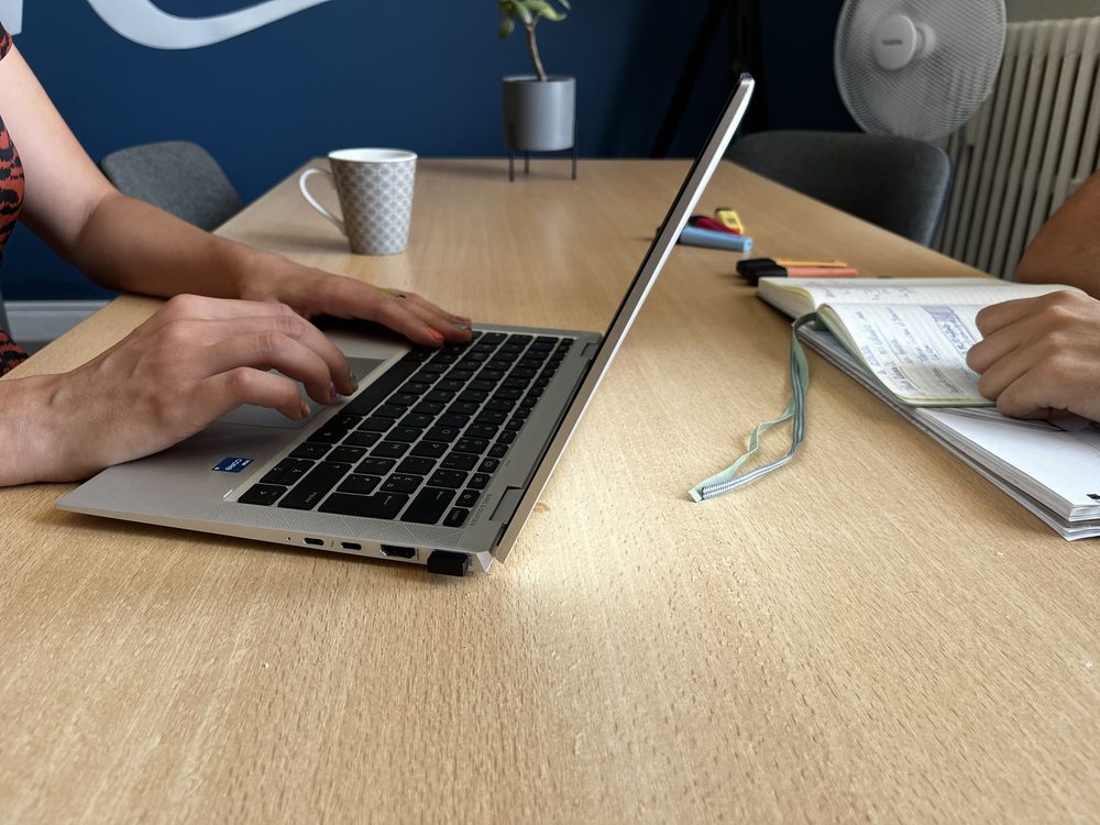 Une main sur le clavier d'un ordinateur portable, un carnet de notes annoté ouvert à côté.
